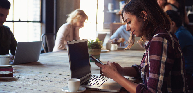 woman in a coffee shop looking at her phone in front of her laptop computer